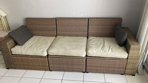 Ein Rattansofa mit Kissen, ein kleiner Tisch mit einem roten Gegenstand und ein Fliesenboden.