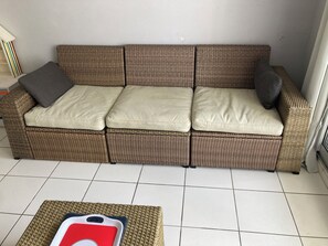 A wicker sofa with cushions, a small table with a red object, and a tiled floor.