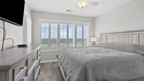 A bedroom with a large bed, a dresser, a ceiling fan, and a view of the ocean through the windows. - Oceanfront views, amenities, all new luxury accommodations in popular Cherry Grove (North Myrtle Beach)