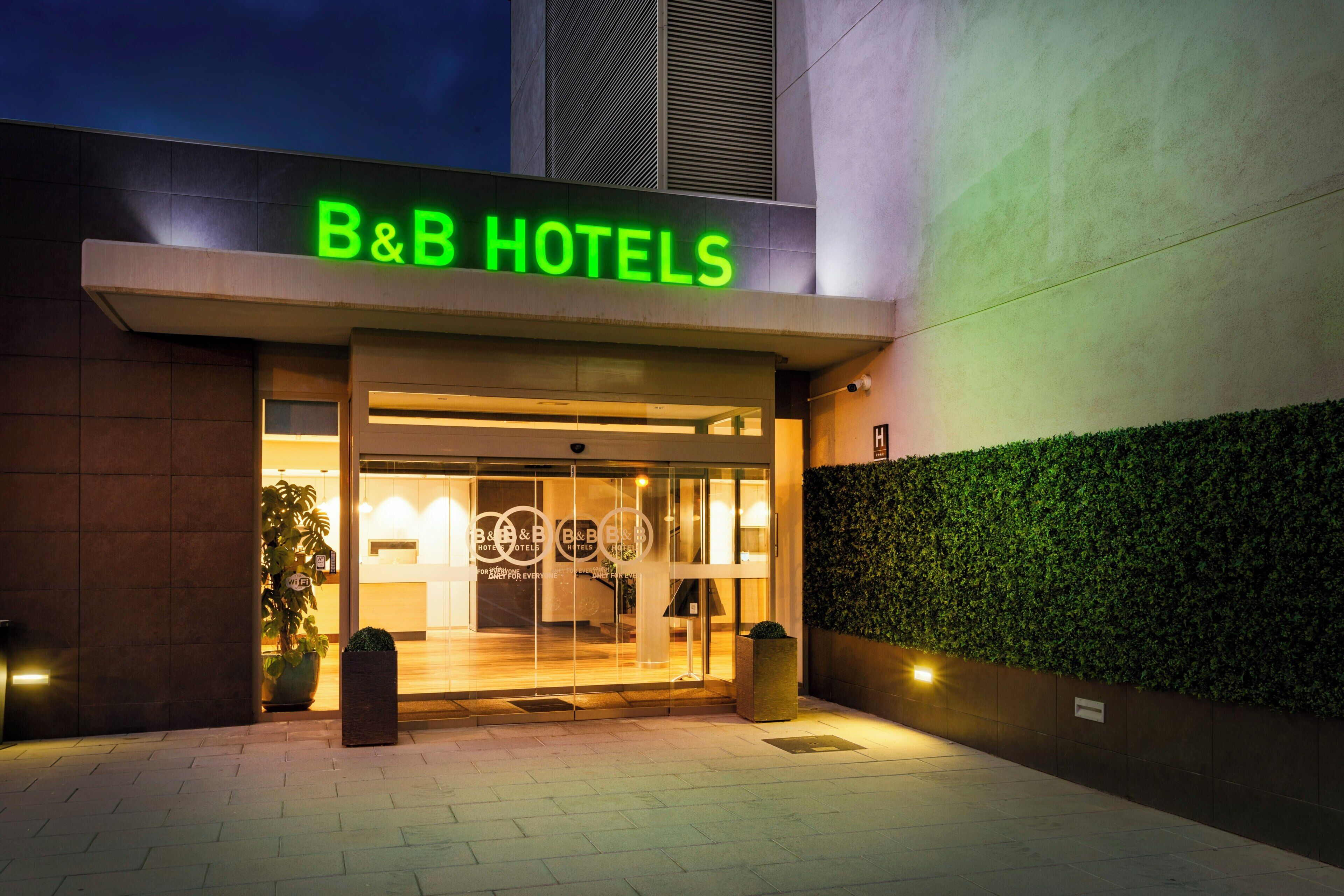 Das moderne Äußere eines B&B Hotels, inklusive Empfangsbereich und einem grünen Heckenstreifen.