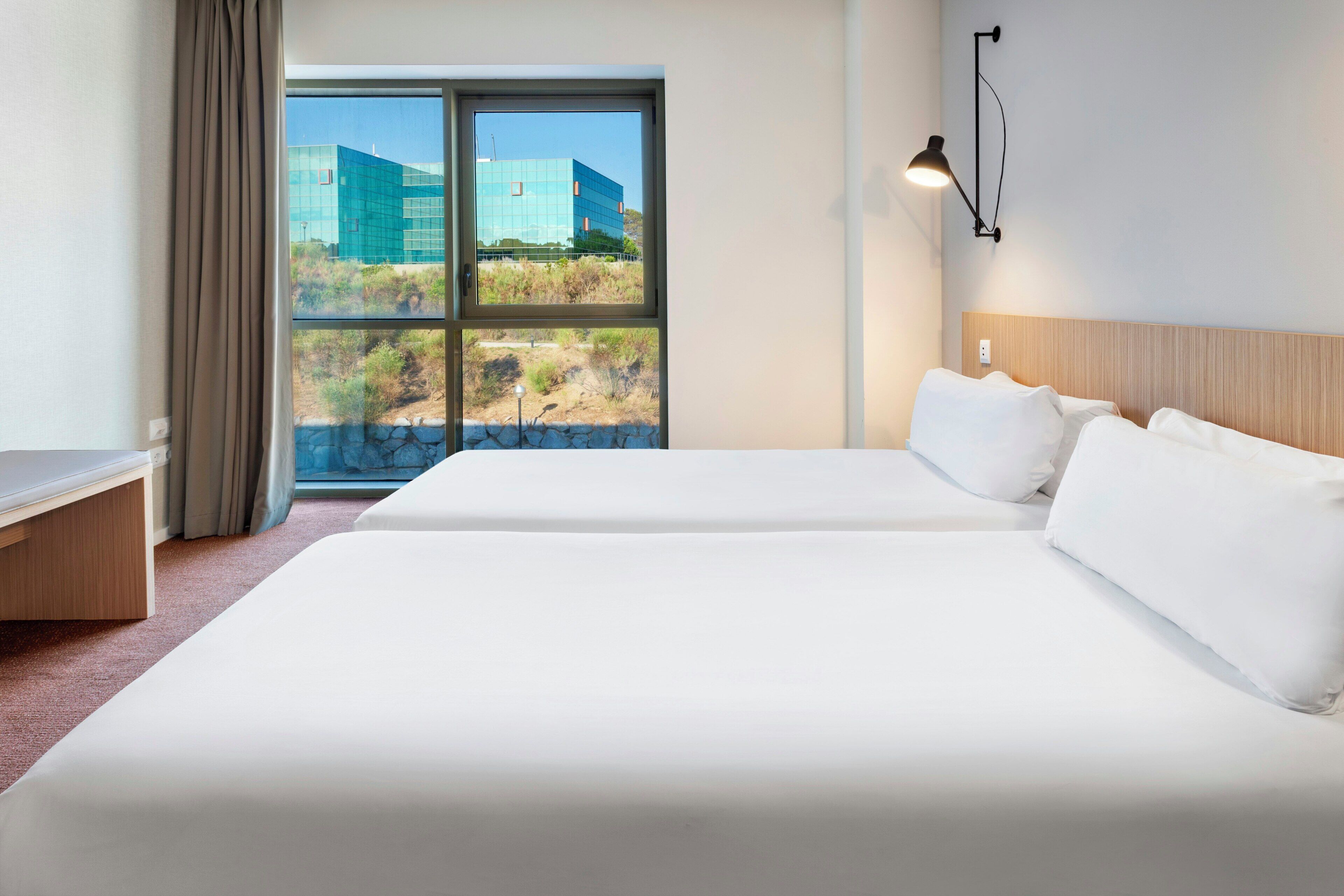 A hotel room with two beds, a desk, and a large window offering a view of a modern building and landscape.