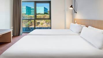 A hotel room with two beds, a desk, and a large window offering a view of a modern building and landscape.