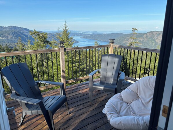 A wooden deck with two Adirondack chairs and a white cushioned seat, overlooking a mountainous landscape and a body of water. - Stunning view above the Malahat (Shawnigan lake)