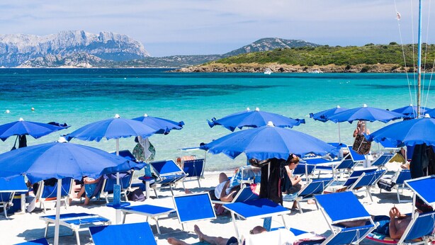 Una playa con hileras de sombrillas azules y tumbonas, un mar azul cristalino y un paisaje montañoso al fondo.