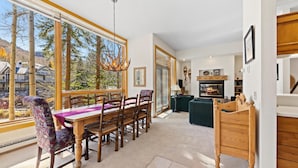 A dining area with a wooden table and chairs, a chandelier, and a view of trees and buildings through large windows. - Cozy Condo: Gore Creek Views & Grand Hyatt Perks (Vai)