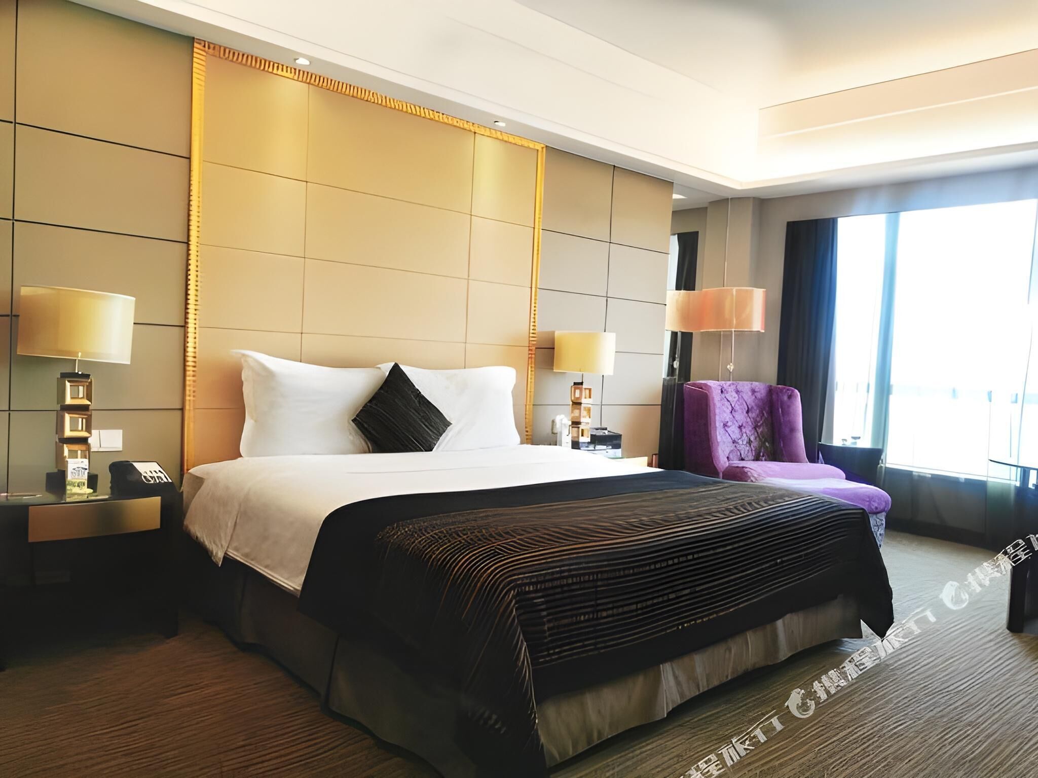 A hotel room with a large bed, two bedside tables, a purple armchair, and a large window.