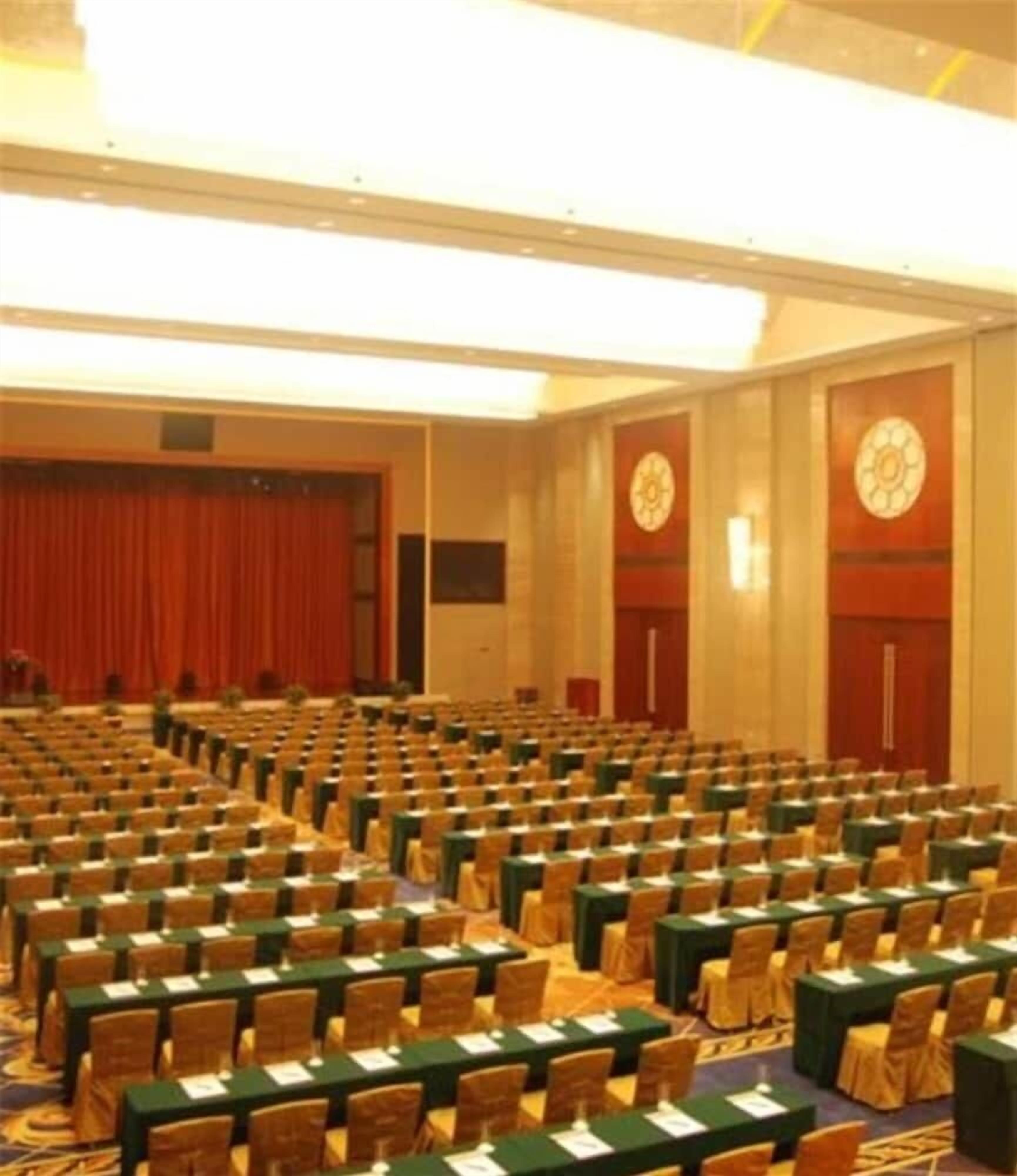 A conference room with rows of tables and chairs, a stage with curtains, and a clock on the wall.
