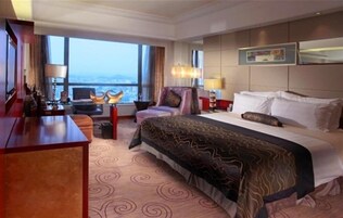 Habitación de hotel con una cama grande, un escritorio, una silla y vistas de la ciudad a través de amplios ventanales.