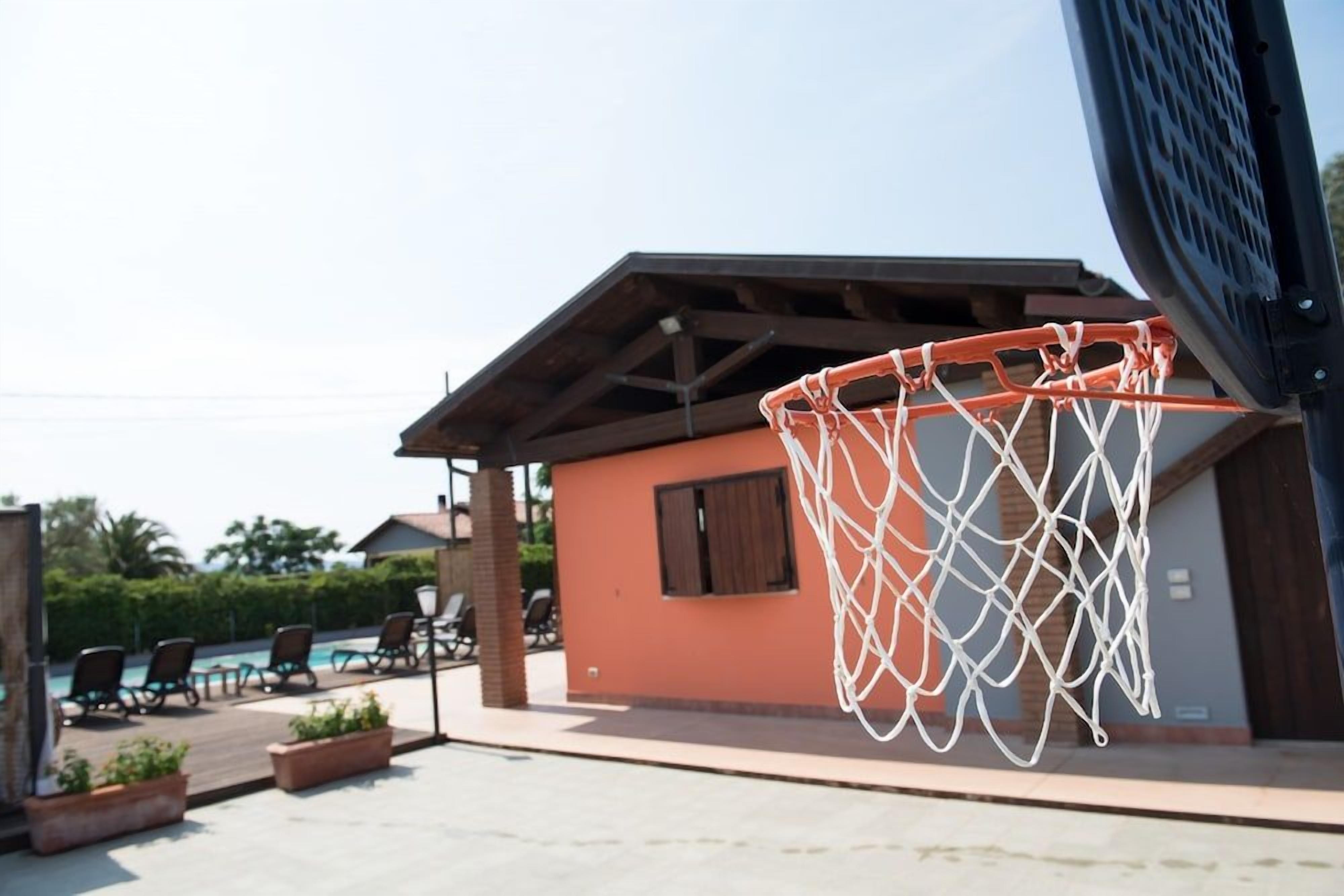 Un panier de basketball installé à l’extérieur, fixé à un bâtiment doté d’un toit, un espace piscine avec des chaises longues et un ciel dégagé.
