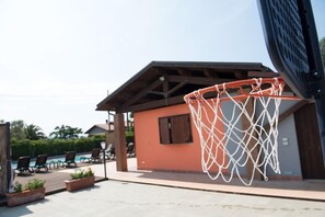 An outdoor basketball hoop attached to a building with a roof, a pool area with lounge chairs, and a clear sky. - Agriturismo Le Due Ruote (Grosseto)