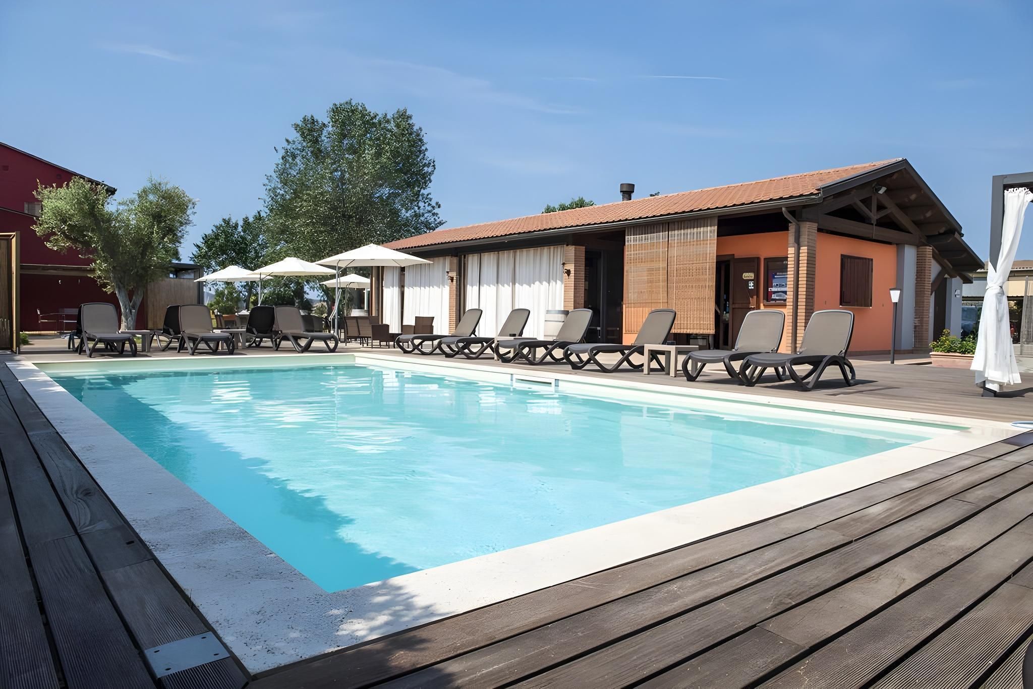 Una piscina con sillas de descanso y sombrillas, rodeada por una terraza de madera y un edificio de techo rojo.