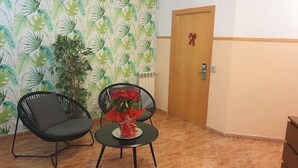 A room with two black chairs, a small round table with a red flower arrangement, a wooden door with a red ribbon, and a patterned wallpaper. - Rio de Castro (Barcelona)