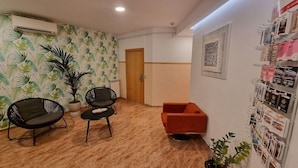 A room with a wooden floor, two black chairs, a red sofa, a potted plant, and a wall with a patterned wallpaper and a framed artwork. - Rio de Castro (Barcelona)