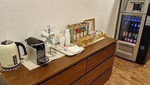 A hotel reception area with a coffee maker, a kettle, a refrigerator stocked with beverages, and a coffee station with cups and condiments. - Rio de Castro (Barcelona)