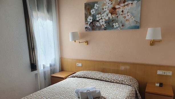 A hotel room with a bed, two bedside tables, a nightstand, a painting of flowers, and a window with curtains. - Rio de Castro (Barcelona)