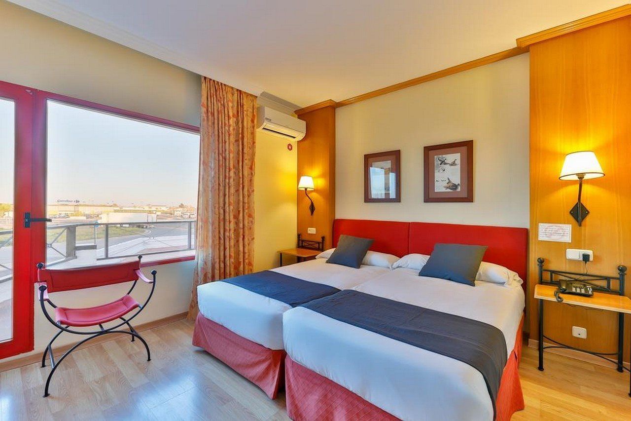 A hotel room with two beds, a bench, a window with a view, and a wall-mounted air conditioner.