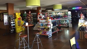 A brightly lit indoor shopping area with a variety of stores, including a visible kiosk with a yellow bear mascot and a section with handbags and accessories. - El Cruce (Manzanares)