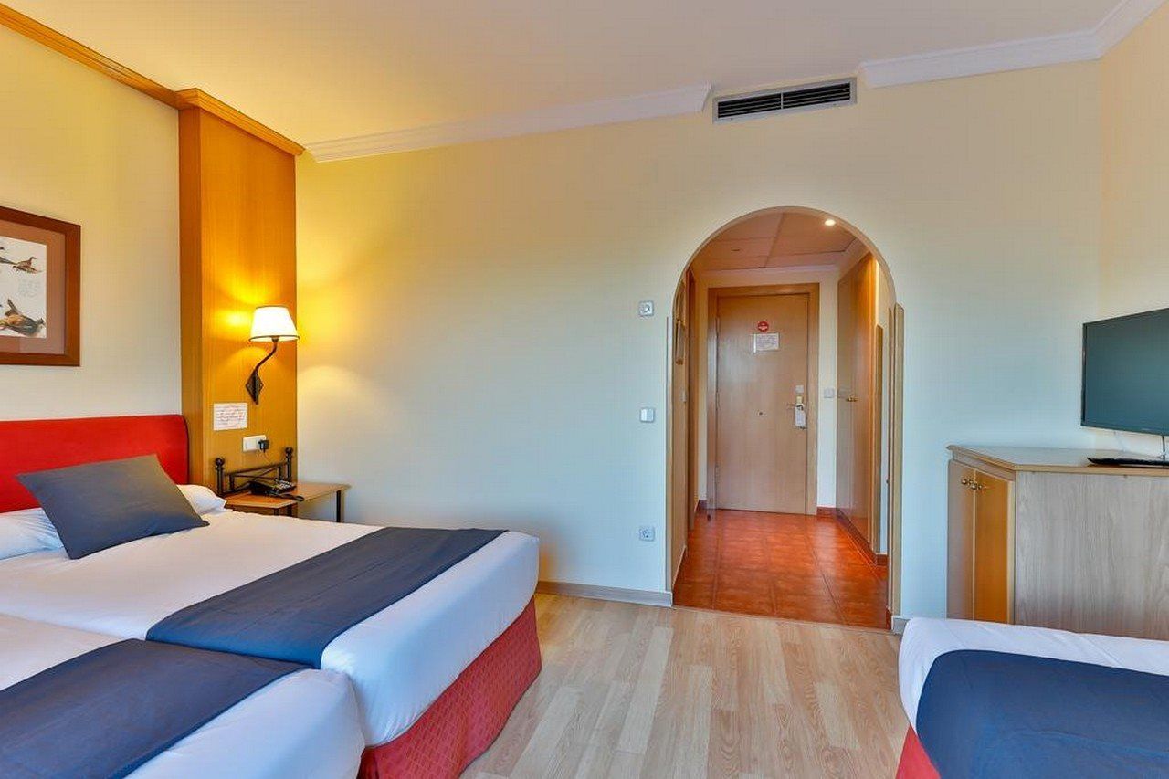 Habitación de hotel con dos camas, un escritorio, un televisor y un pasillo que conduce a otra habitación.