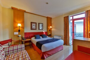 Habitación de hotel con cama, dos mesitas de noche, una silla, un escritorio y un ventanal con cortinas.