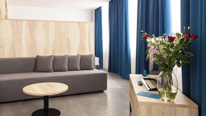 A modern hotel room with a grey sofa, a round wooden table, and a vase with flowers on a wooden side table.