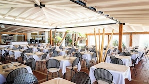 A spacious outdoor dining area with multiple tables and chairs, covered by a large white canopy, and surrounded by wooden beams and potted plants.