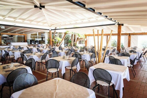 A spacious outdoor dining area with multiple tables and chairs, covered by a large white canopy, and surrounded by wooden beams and potted plants. - Baia Dei Mulini (Erice)