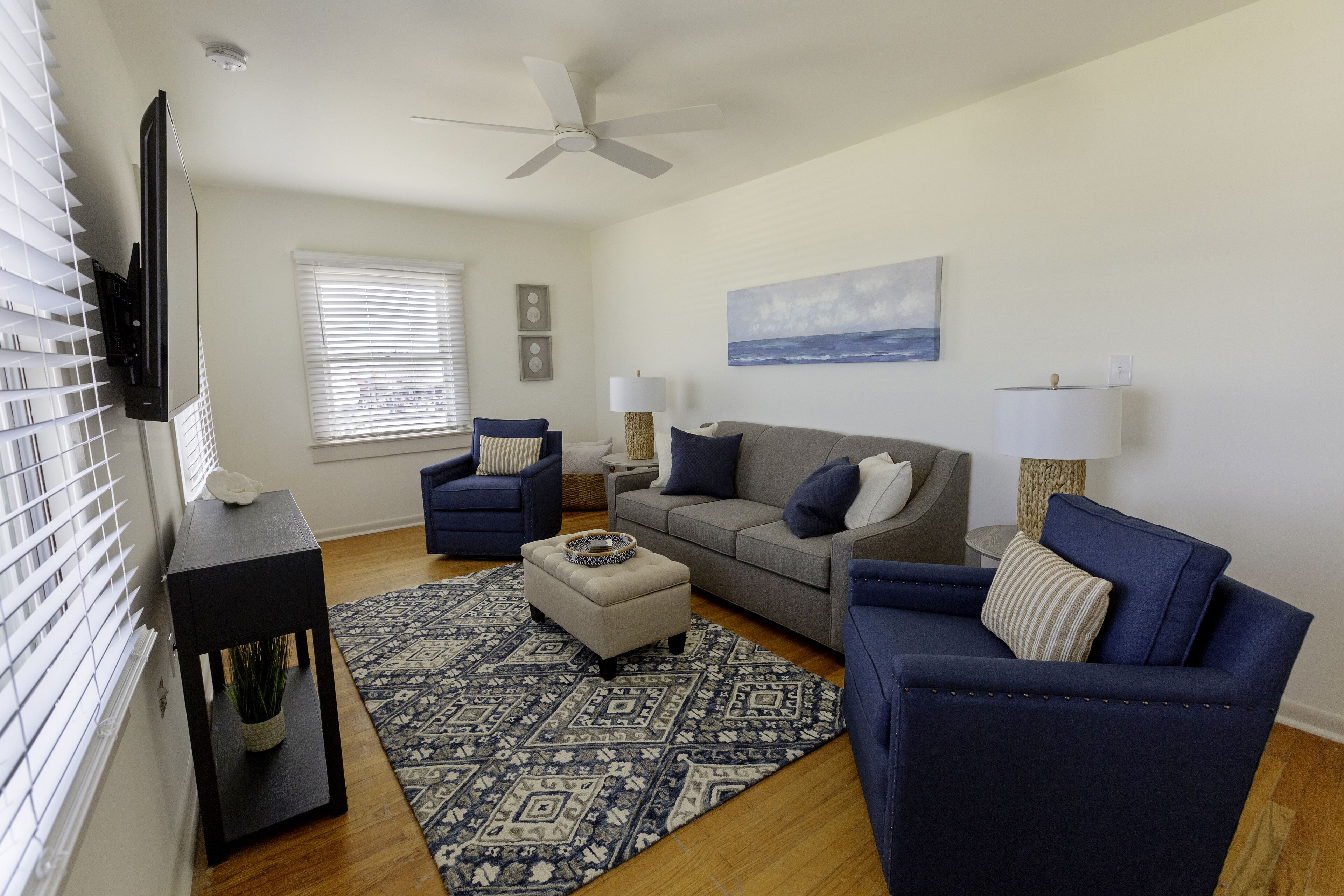 Una sala de estar con un sofá gris, un sillón azul, un otoman blanco y un televisor montado en la pared.