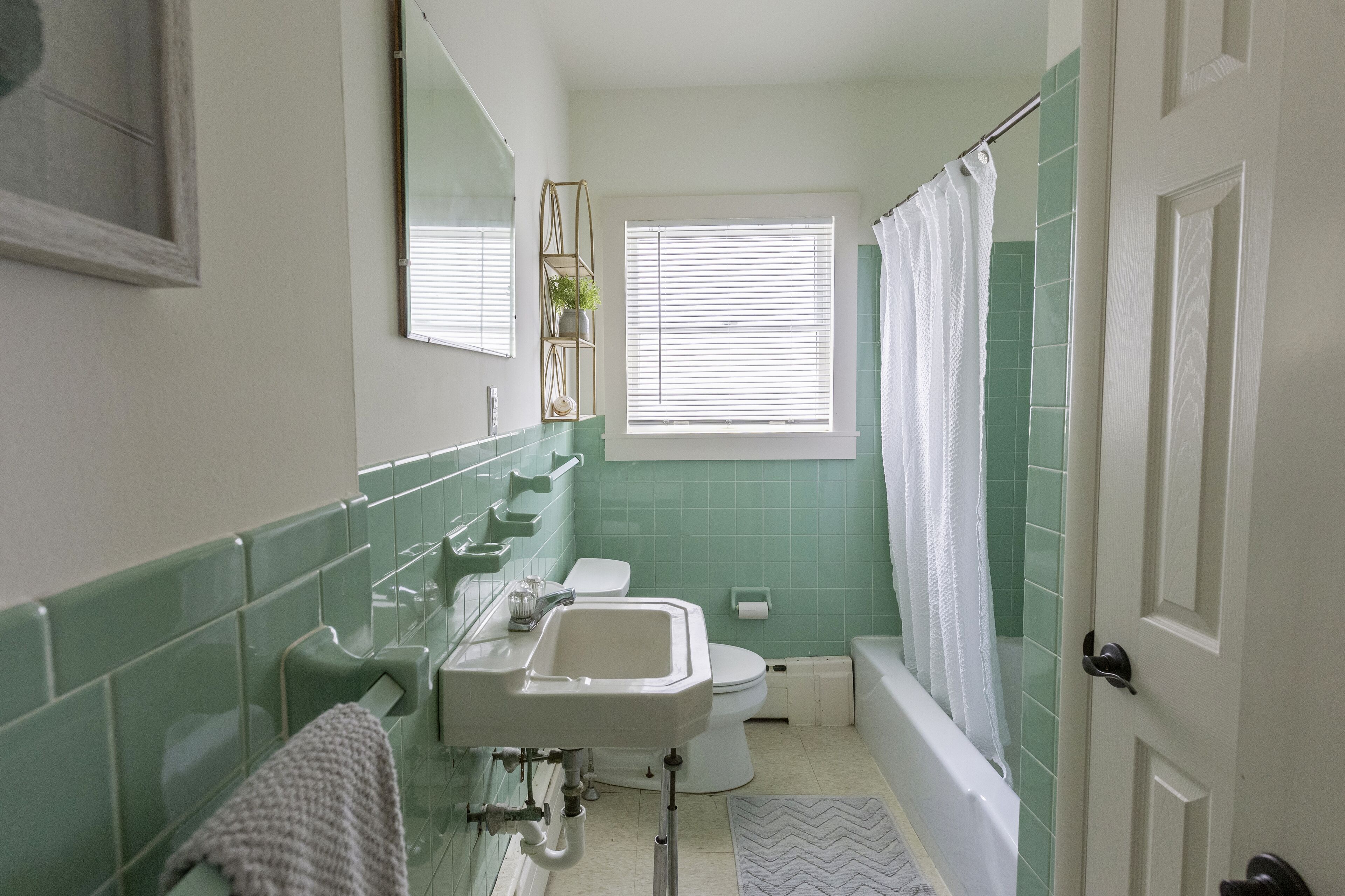 A bathroom with a white sink, toilet, and bathtub, green tiles, and a window with blinds.