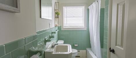 A bathroom with a white sink, toilet, and bathtub, green tiles, and a window with blinds.
