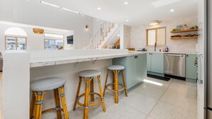 A modern kitchen with a marble countertop, three bamboo stools, a stainless steel dishwasher, and a staircase leading to another level. - STUNNING! Ocean Views!! Lux Remodeled Modern Beach House. +PELETON . (Oxnard)