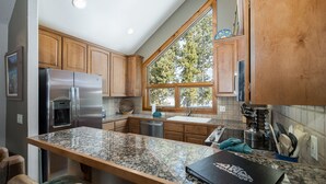 A kitchen with wooden cabinets, a granite countertop, a stainless steel refrigerator, a built-in oven, a sink, and a window with a view of trees.