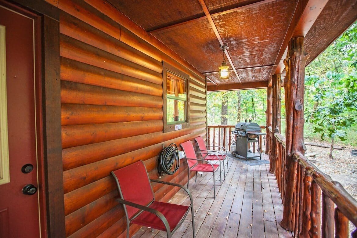 A wooden cabin with a porch, red chairs, and a grill.