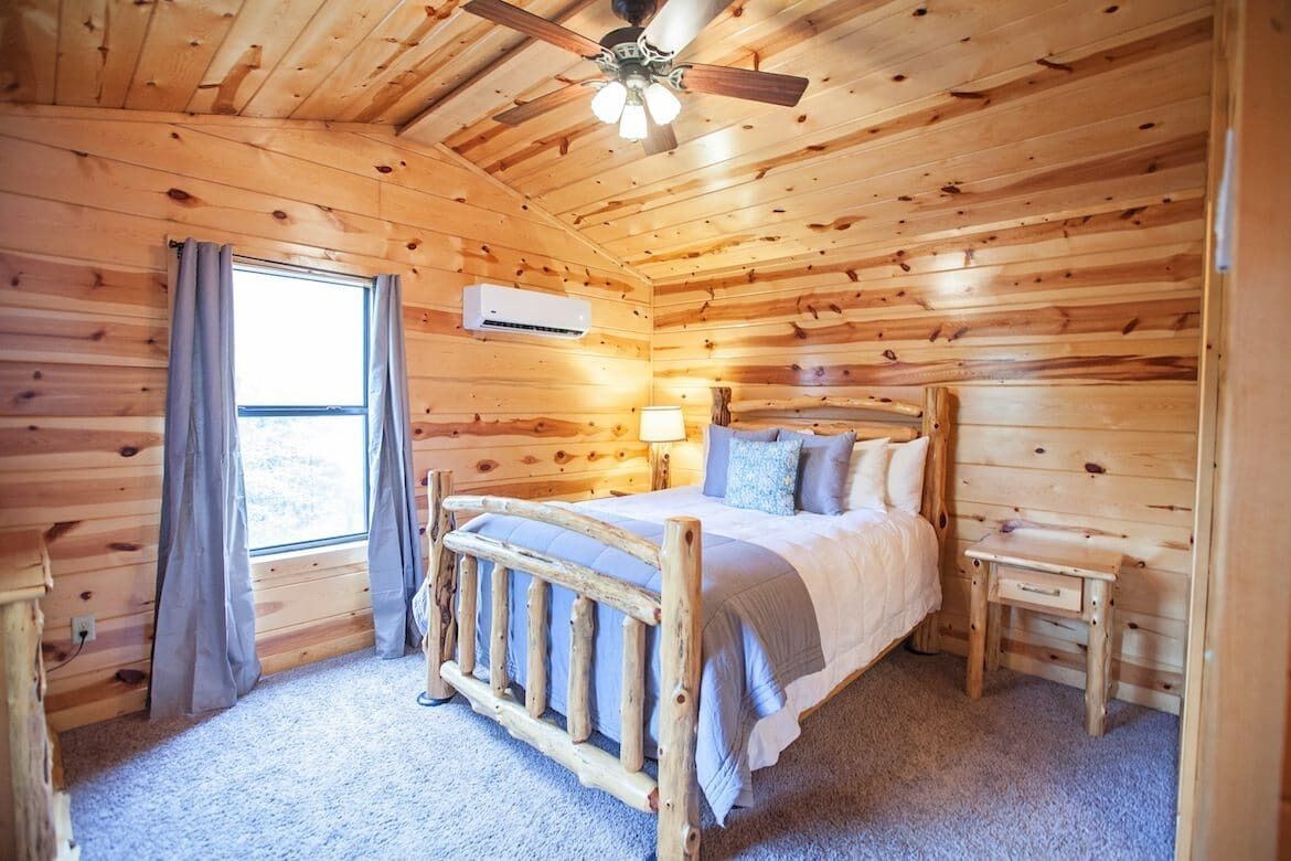 Een slaapkamer in een houten chalet met een bed, nachtkastje, plafondventilator en raam met gordijnen.