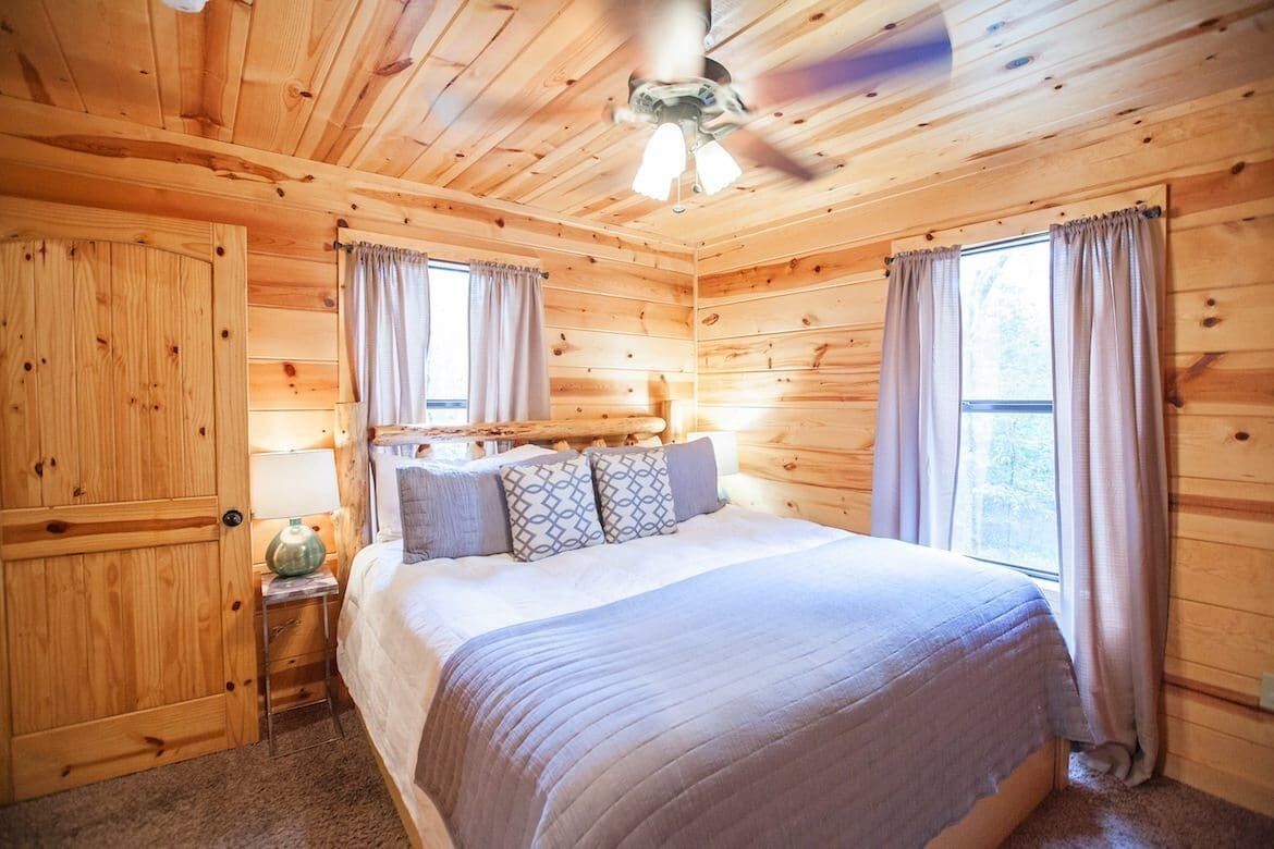 A wooden cabin bedroom with a bed, curtains, a ceiling fan, and a nightstand with a lamp.