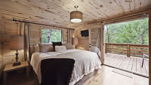 Une chambre de style cabane en bois, avec un grand lit, une table de chevet avec une lampe, un téléviseur fixé au mur et un balcon doté d’une balustrade en bois.