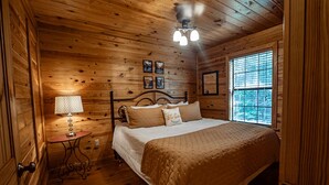 Ein Schlafzimmer einer Holzhütte mit einem Bett, einem kleinen Tisch mit Lampe und einem Fenster mit Jalousien.