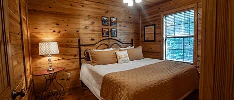 A wooden cabin bedroom with a bed, a small table with a lamp, and a window with blinds.