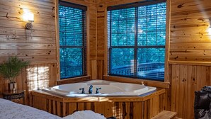 A wooden cabin interior with a large bathtub, two windows with blinds, and a small table with a plant.