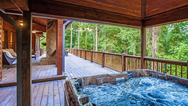 A wooden deck with a hot tub, stone pillar, and a view of the forest. - Sugarberry Mountain Lodge-4 BR-Timber Creek Trails (Broken Bow)