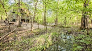Une cabane dotée d’une terrasse et de meubles de jardin, entourée d’arbres et bordée par un ruisseau.