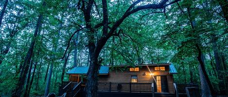 A cabin in a forest with a wooden exterior, a deck, and a staircase leading to the entrance.