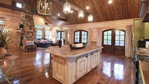 A spacious log cabin interior with a large central island, stone fireplace, and multiple windows. - Maison de Crique- Studio -Timber Creek Trails (Broken Bow)