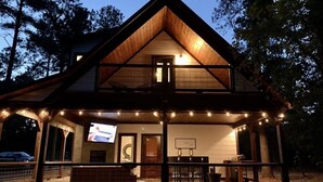 Una casa de madera de dos pisos con balcón, una pantalla de televisión y luces colgantes.