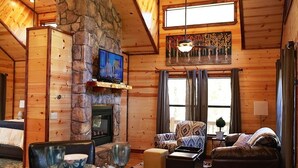 L’intérieur d’un chalet confortable, avec des murs en rondins de bois, une cheminée en pierre, un téléviseur fixé au mur et un coin salon accueillant, comprenant un canapé et des fauteuils.