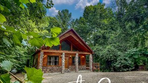 Une cabane en bois, posée sur des fondations en pierre, entourée d’une végétation luxuriante et surplombée d’un ciel bleu azur.