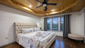 A bedroom with a large bed, a ceiling fan, wooden flooring, and a window with curtains.