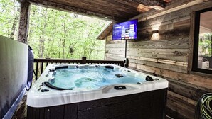 A wooden cabin with a hot tub, a mounted TV displaying streaming services, and a view of the surrounding forest. - Cherokee Rose- 1BR-Northern Hills (Broken Bow)