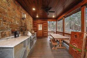 L’intérieur d’une cabane avec un mur en pierre, un plafond en bois, un barbecue, une table en bois et un banc.