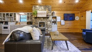 A cozy cabin interior with a stone fireplace, leather sofa, wooden floors, and a flat-screen TV mounted on the wall. - Summer Brook Studio-Creekside-Timber Creek Trails (Broken Bow)