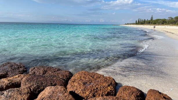 Ein Strand mit klarem türkisblauem Wasser, einem sandigen Ufer und großen Felsen im Vordergrund.